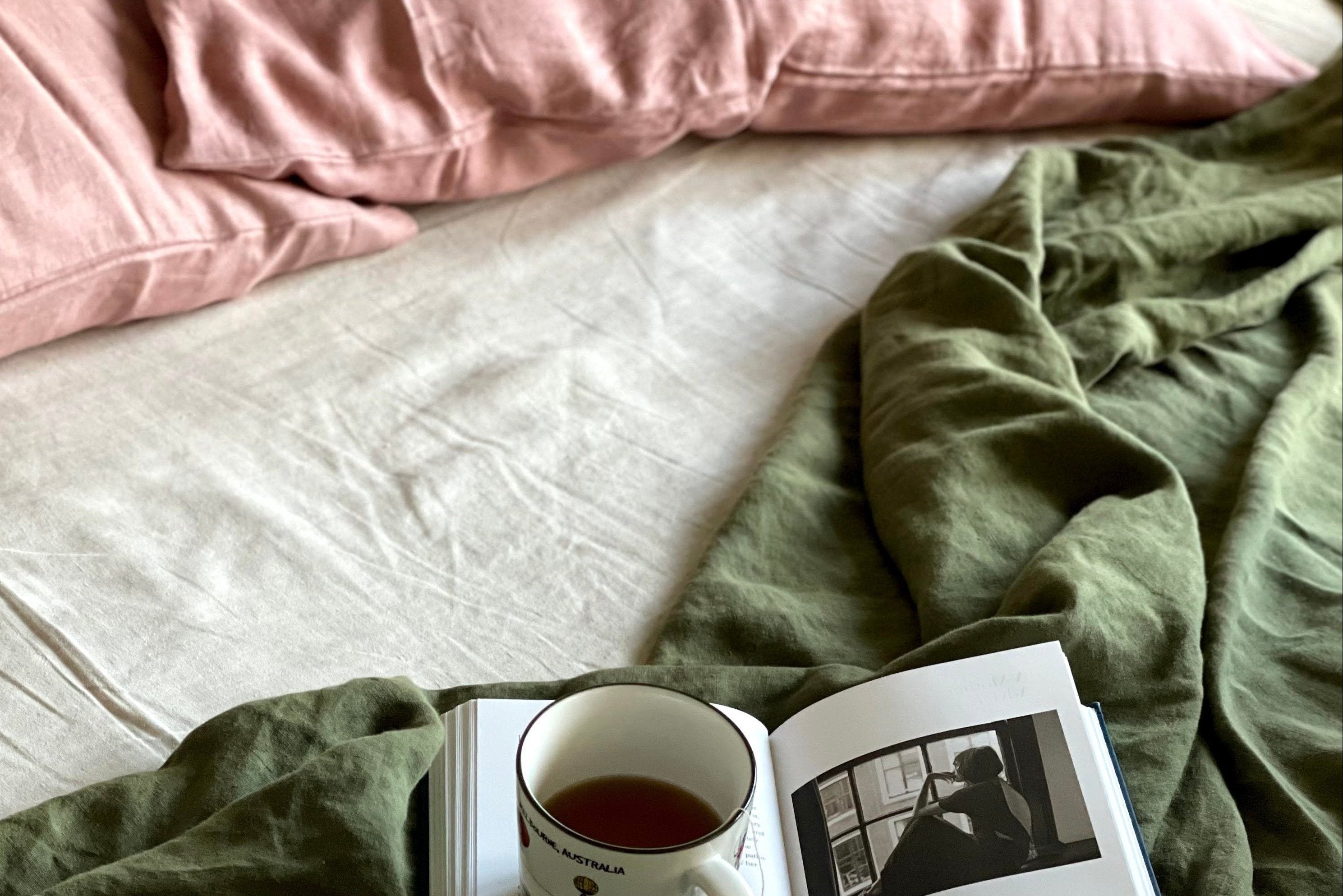 Book with a cup of tea on a bed with pink pillows and green sheet.